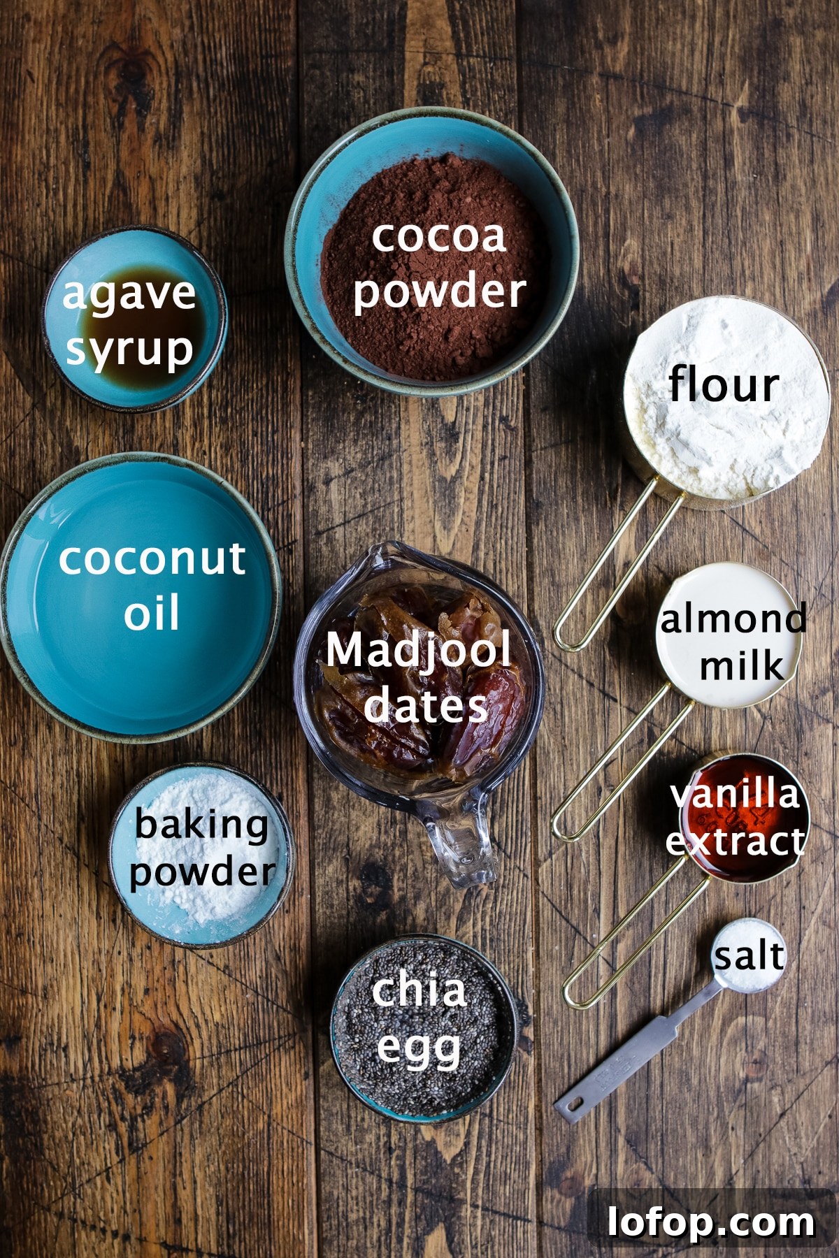 Various ingredients for a vegan brownie layer, such as flour, cocoa powder, agave juice, Medjool dates, chia egg, baking powder, coconut oil, vanilla extract, and almond milk, organized on a kitchen counter.