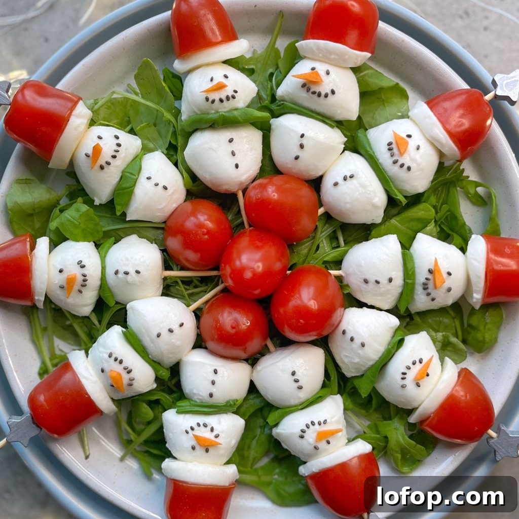 A plate of salad arranged with mozzarella and tomato snowmen, each with black dots for eyes and mouth, and a carrot nose, on a bed of greens—fun bites that make healthy food recipes festive. Cherry tomatoes form Santa hats at the center of the dish.