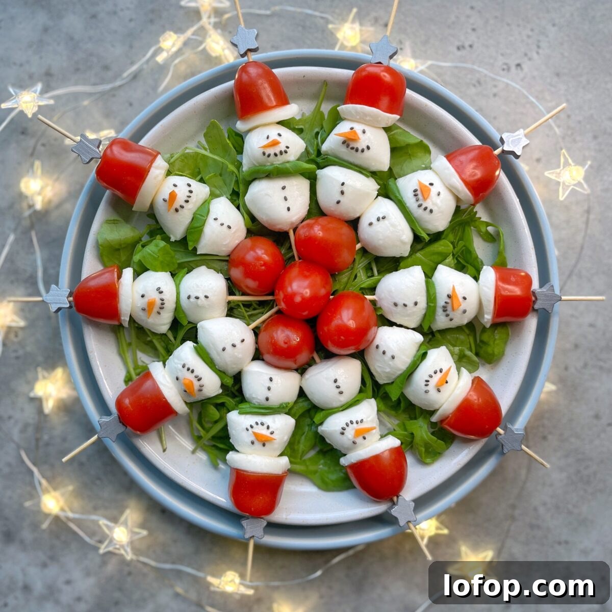 A plate of salad with skewers arranged in a circle, each decorated to look like snowmen using mozzarella, tomatoes, and arugula. A festive string of lights surrounds the plate.