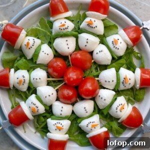 A plate of salad arranged with mozzarella and tomato snowmen, each with black dots for eyes and mouth, and a carrot nose, on a bed of greens—fun bites that make healthy food recipes festive. Cherry tomatoes form Santa hats at the center of the dish.