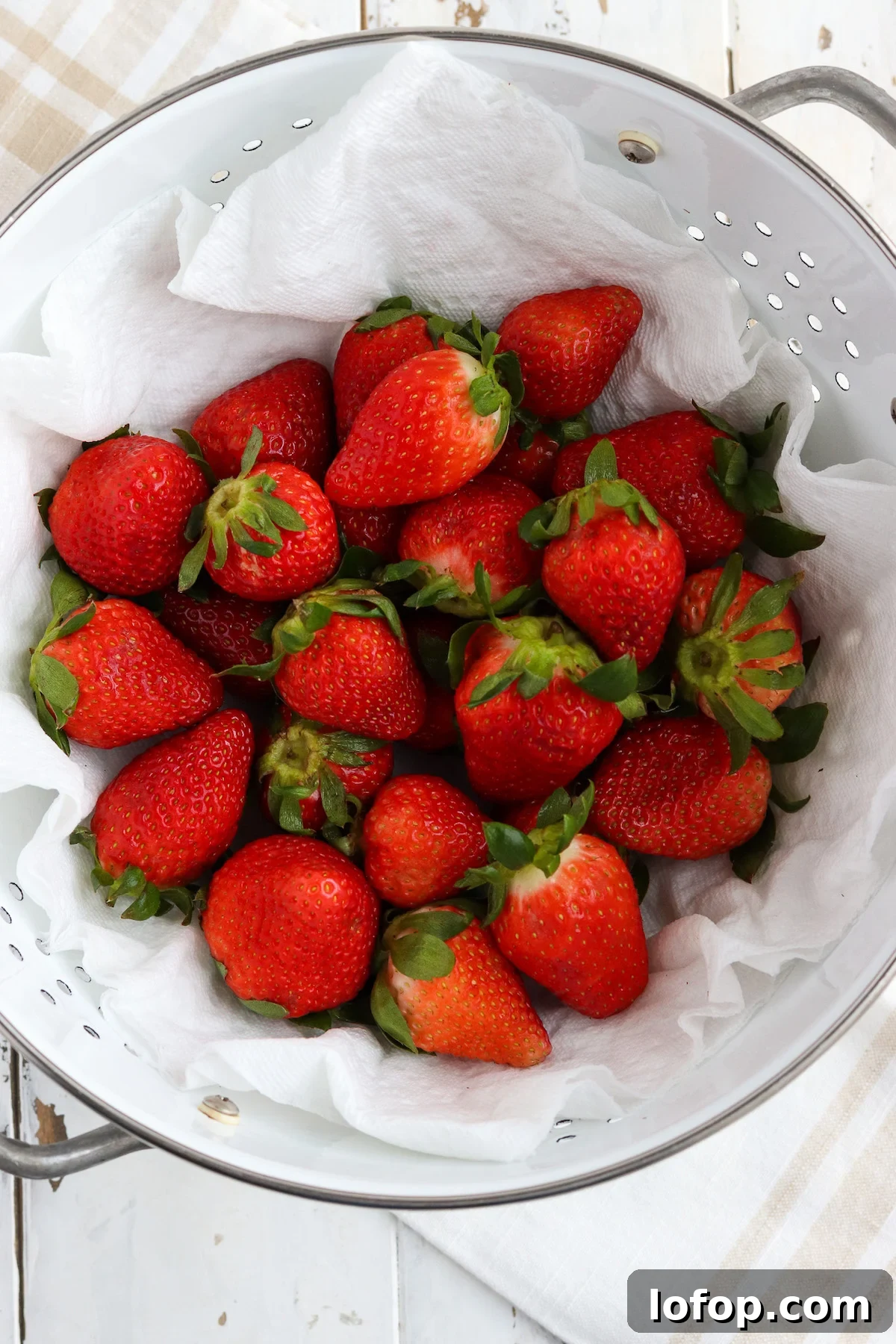 A colander of washed bright red strawberries drying on a paper towel.