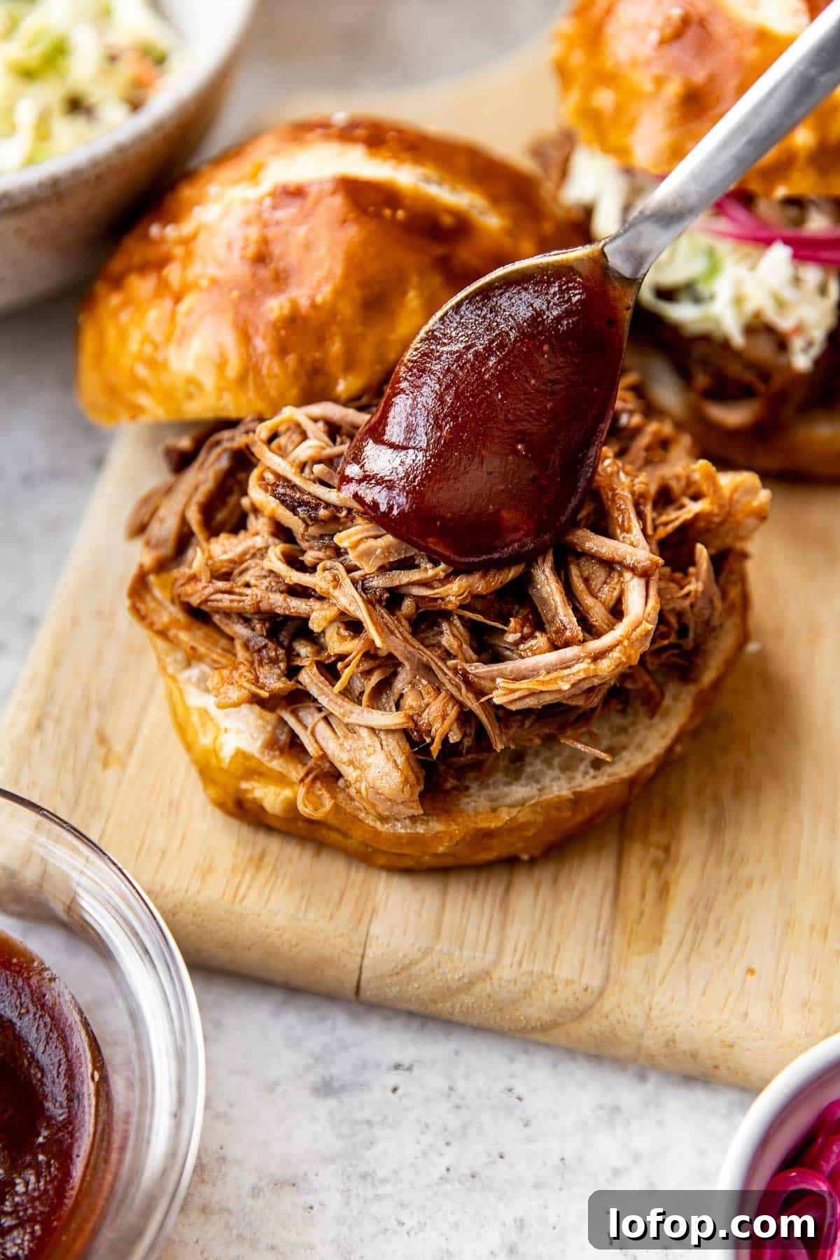 pulled pork on a pretzel bun with bbq sauce being spooned on top of the pork.