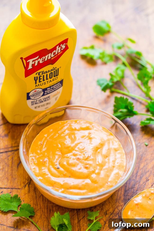 A bottle of French’s Classic Yellow Mustard next to a glass bowl of creamy orange sauce, with cilantro leaves scattered on a wooden surface.