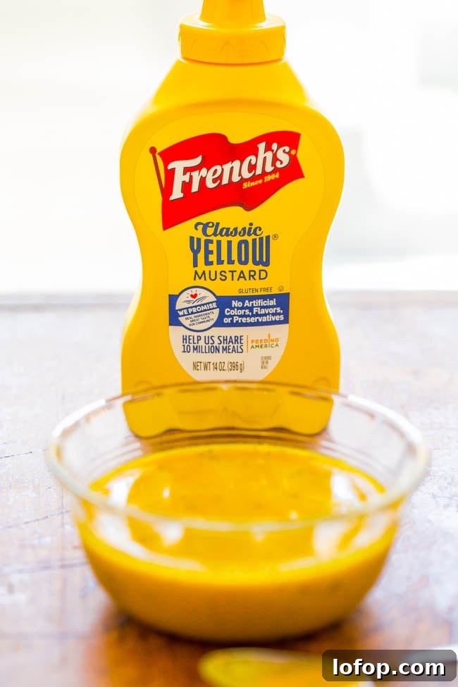 A bottle of French's Classic Yellow Mustard stands behind a glass bowl filled with mustard on a wooden surface.