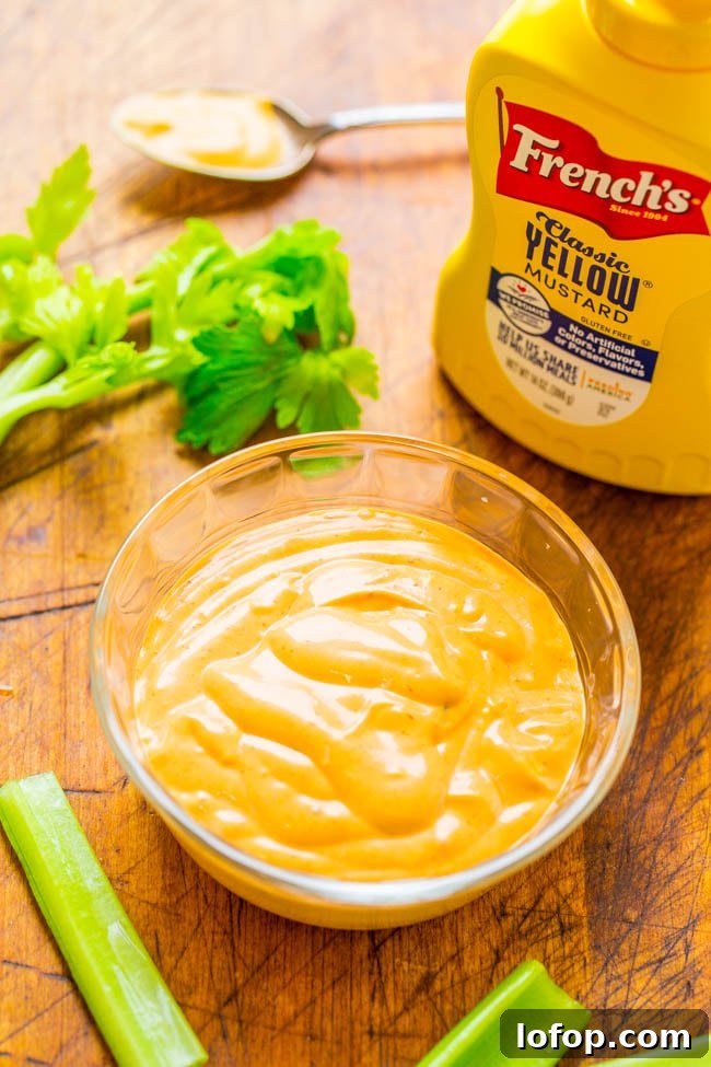 A glass bowl of creamy yellow mustard sauce sits on a wooden surface next to celery stalks, a spoon, and a bottle of French's Classic Yellow Mustard.