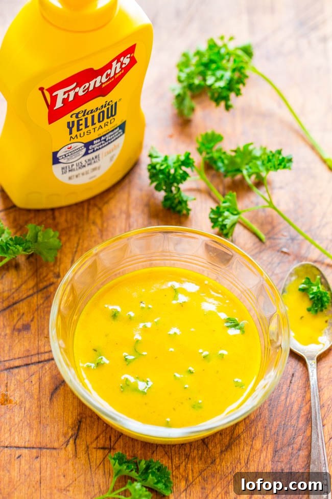 A bowl of yellow mustard sauce garnished with herbs sits on a wooden surface beside a French's mustard bottle, parsley sprigs, and a spoon.