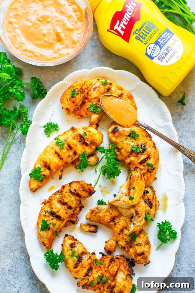 Grilled chicken tenders on a white plate, garnished with parsley, served with a spoonful of sauce. French’s yellow mustard and a bowl of sauce are visible nearby.