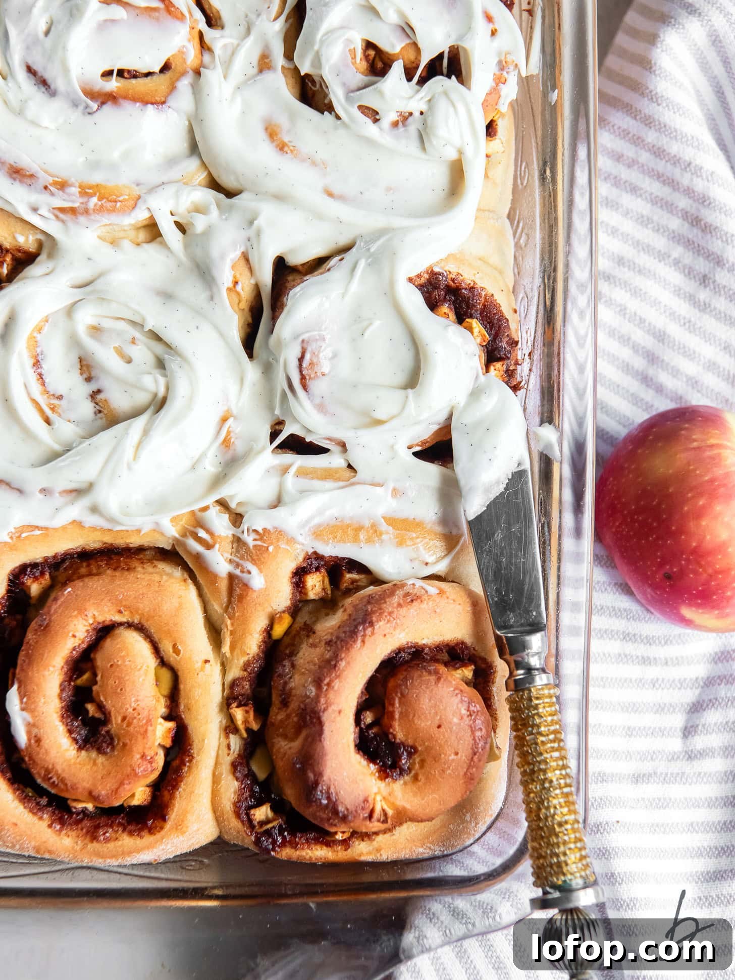 knife spreading cream cheese icing on top of apple cinnamon rolls in a baking dish.