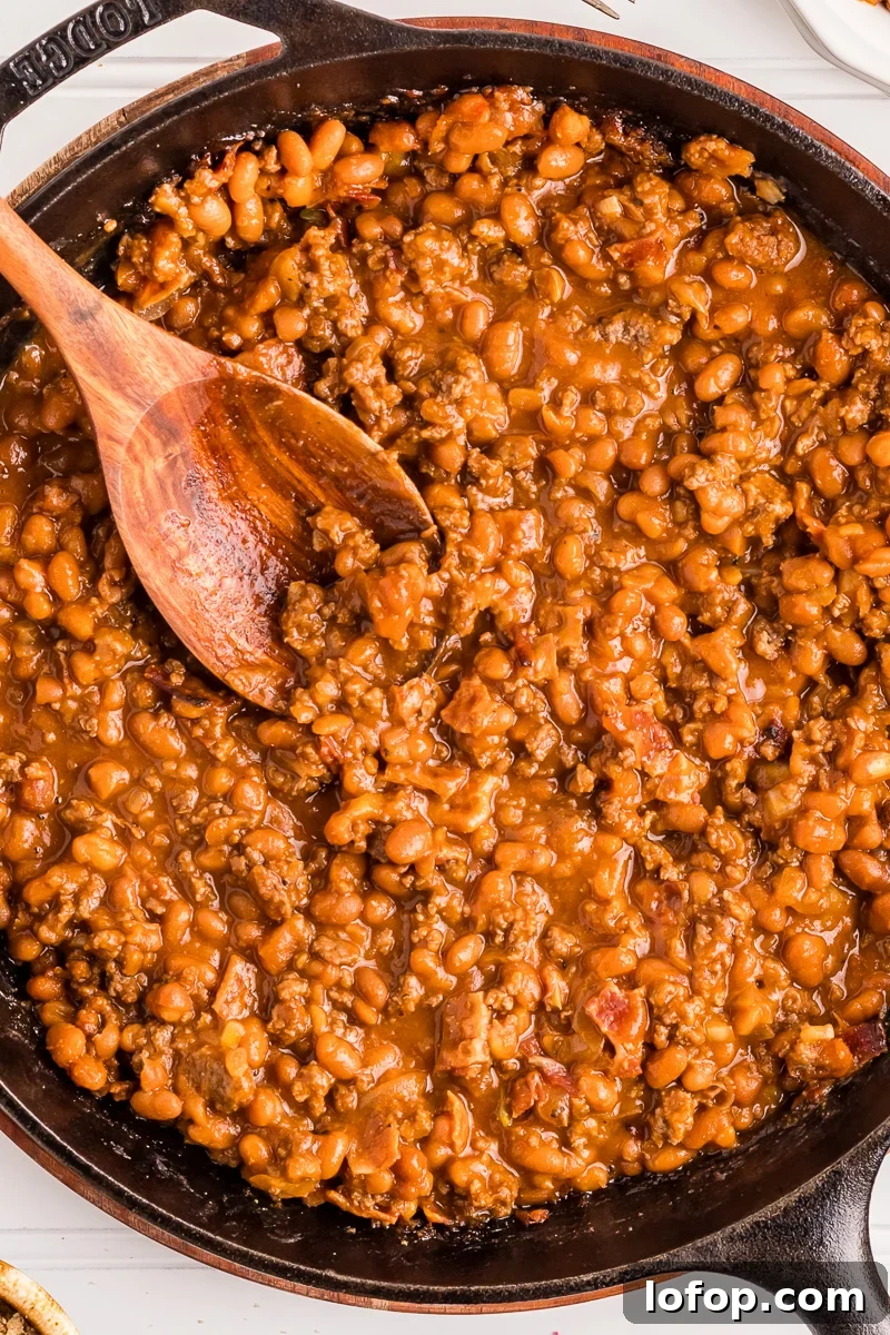 Hearty Baked Beans with Ground Beef — Stovetop, Oven or Slow Cooker 2 Baked beans with ground beef and bacon in cast iron skillet with a spoon.