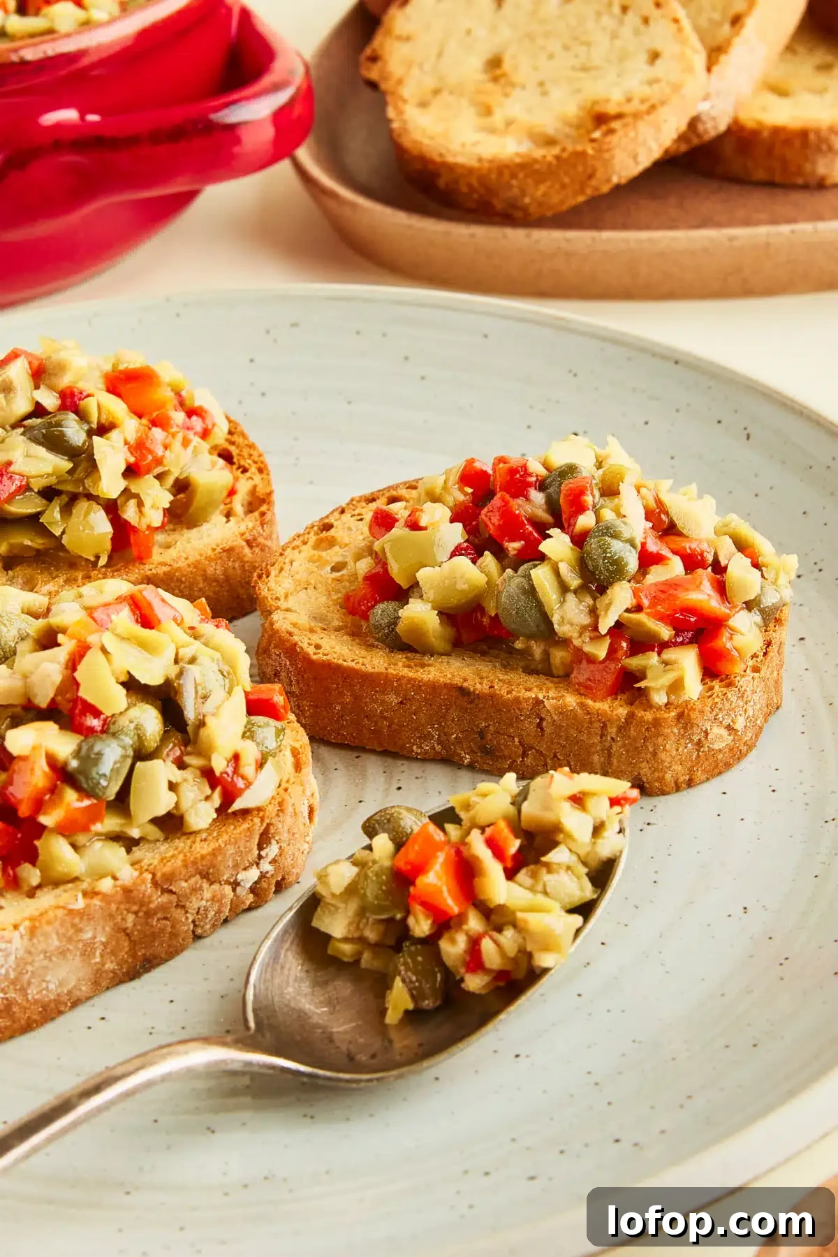 Toasted baguette slices topped with olive tapenade are arranged on a rustic grey serving plate. A spoon filled with tapenade sits on the plate, and a plate of plain toasted bread slices sits on the side.