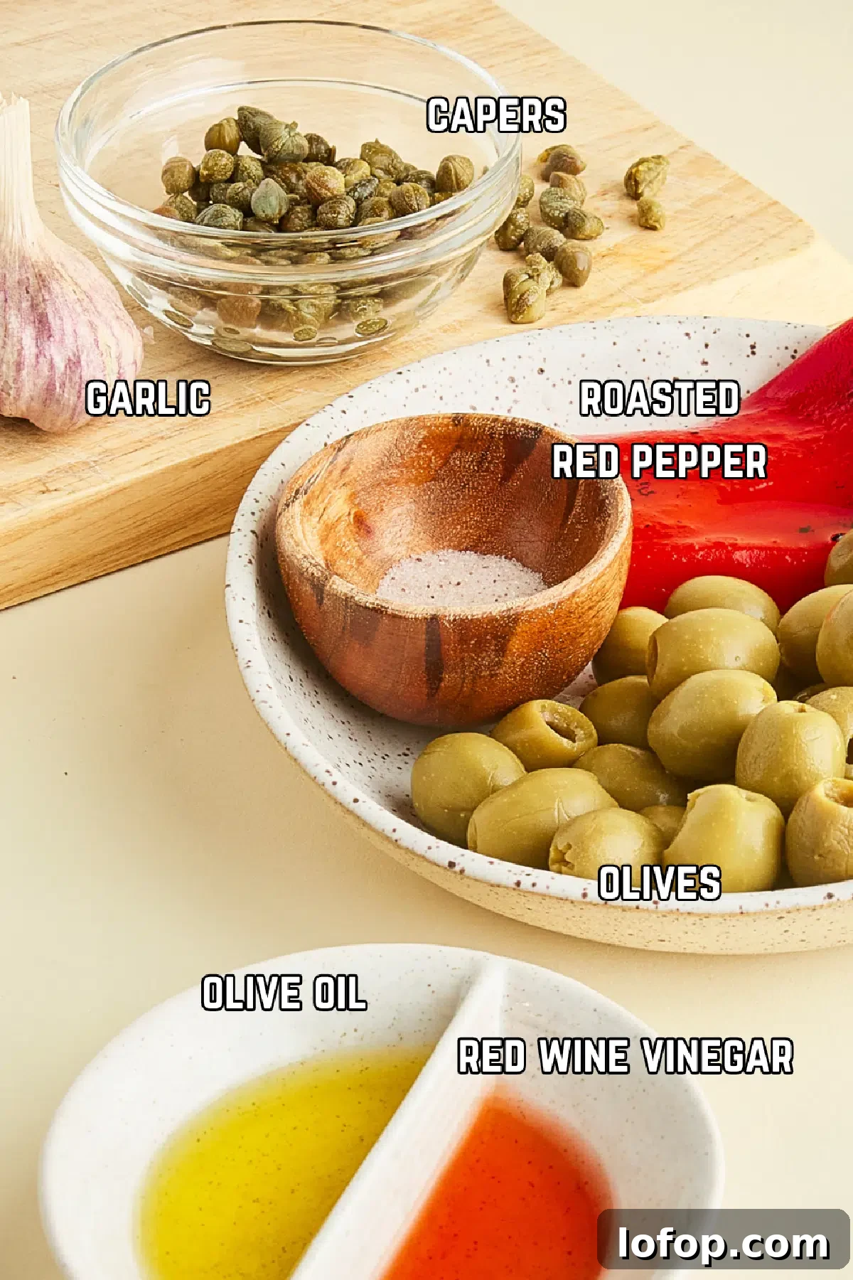 Ingredients to make tapenade sit in various bowls and boards: capers, garlic, roasted red pepper, olives, olive oil, and vinegar.