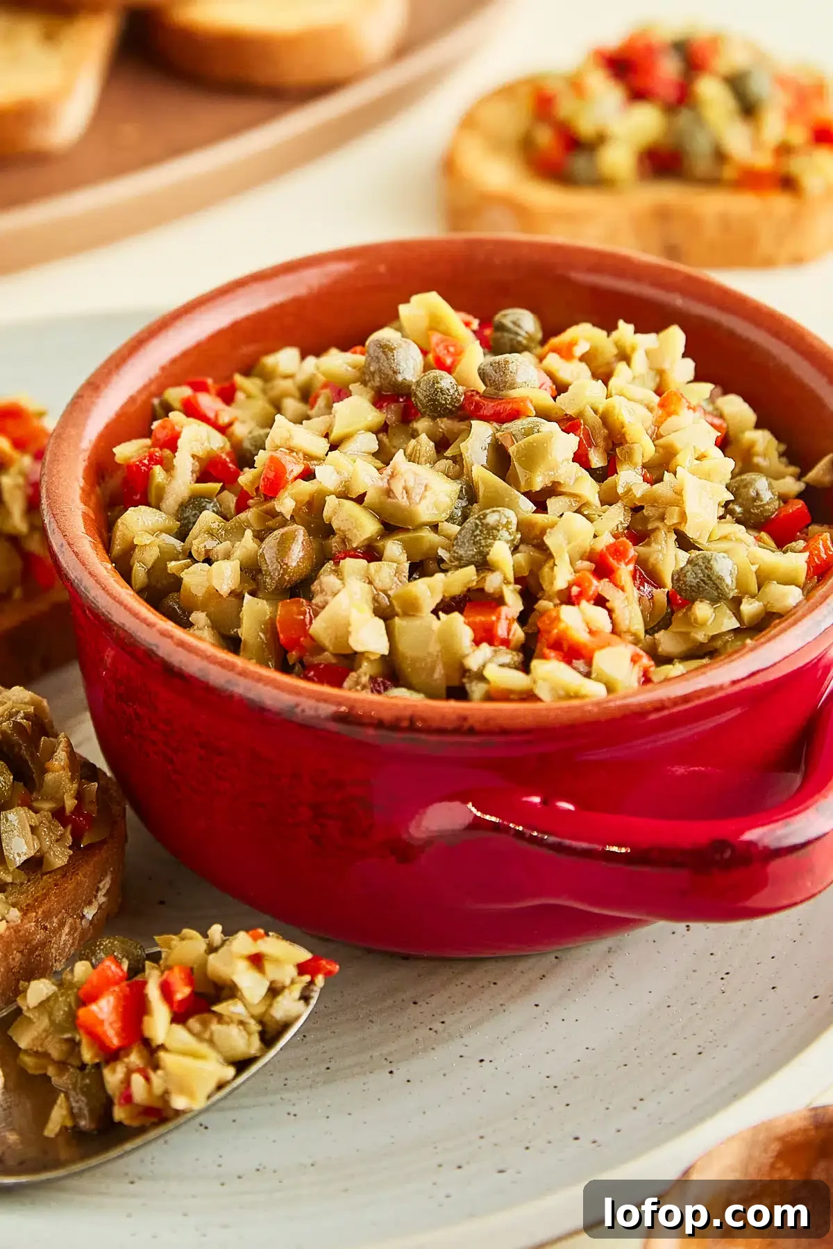 A red bowl of olive tapenade sitting on a rustic grey plate that holds two toasted baguette slices topped with tapenade, and a spoon filled with tapenade. Another plate of plain breads sits on the side.
