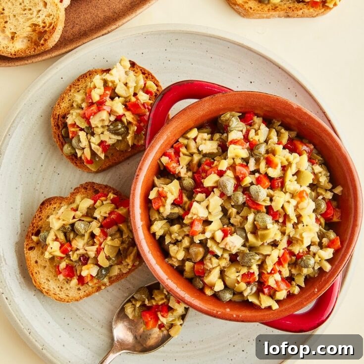 Overhead view of a red bowl of olive tapenade sitting on a rustic grey plate that holds two toasted baguette slices topped with tapenade, and a spoon filled with tapenade. Another plate of plain breads sits on the side.
