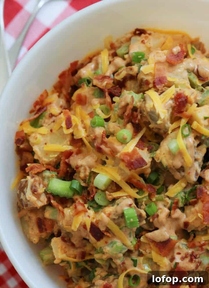 A large white bowl of BBQ potato salad garnished with cheddar cheese, bacon, and green onions on a red and white check gingham cloth.