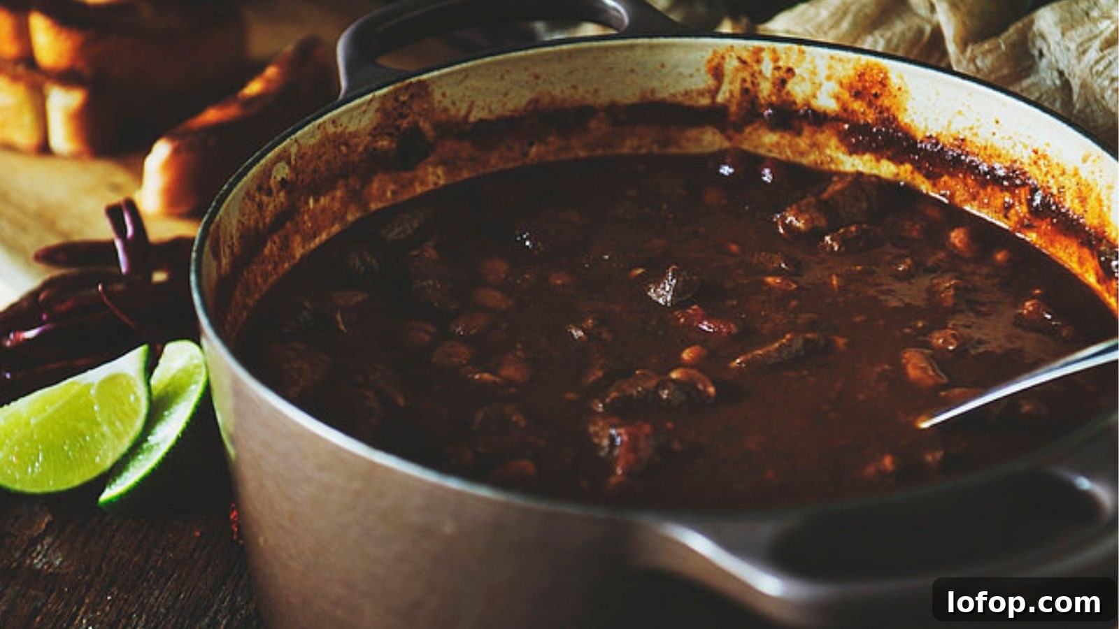 12 Epic Chili Recipes for Game Day Crowd-Pleasing Flavor 2 A pot of rich, dark chili containing beans and meat, with a metallic ladle. Lime wedges and dried chilies are visible in the background.