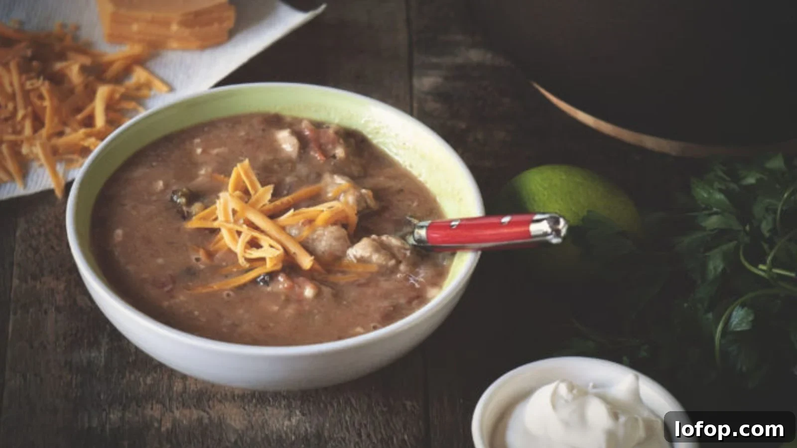 12 Epic Chili Recipes for Game Day Crowd-Pleasing Flavor 11 Bowl of soup with chunks of meat and vegetables, topped with shredded cheese. A red-handled spoon rests in the bowl. Surrounding items include shredded cheese, lime, herbs, and sour cream.