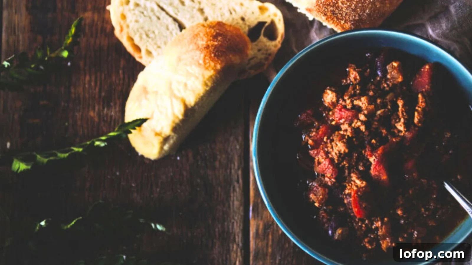 12 Epic Chili Recipes for Game Day Crowd-Pleasing Flavor 12 A bowl of chili, inspired by classic chili recipes, sits on a wooden table with a spoon, accompanied by sliced bread and fresh green leaves.