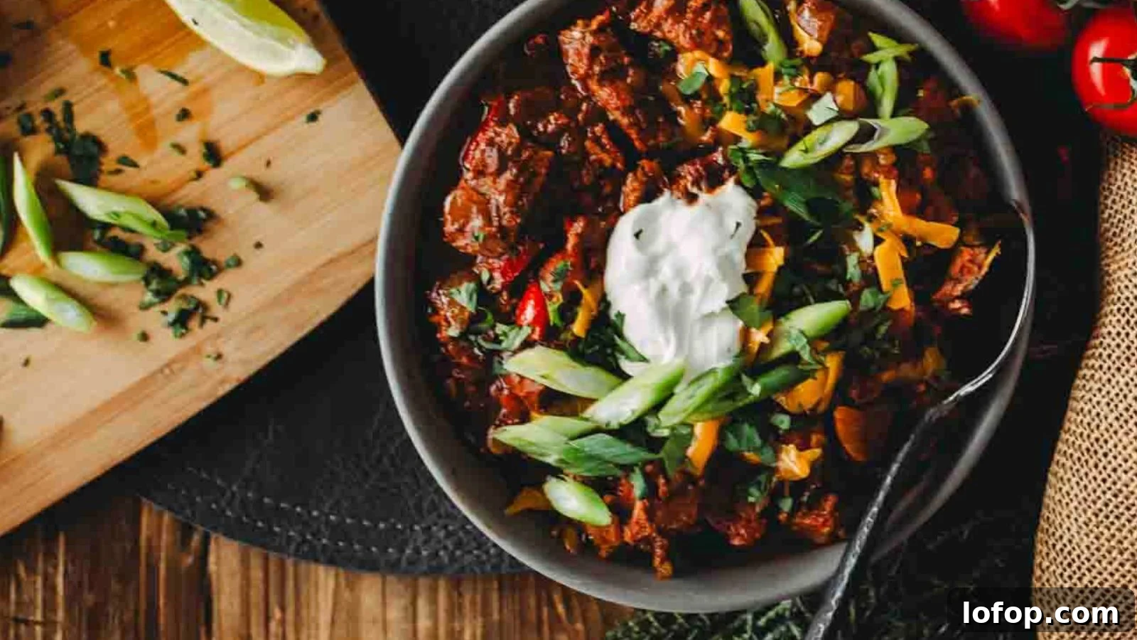 12 Epic Chili Recipes for Game Day Crowd-Pleasing Flavor 3 A bowl of chili topped with sour cream, sliced green onions, and cilantro, with a wooden board of lime wedges and chopped herbs nearby. A spoon rests in the bowl.