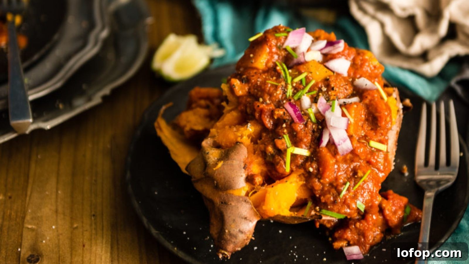 12 Epic Chili Recipes for Game Day Crowd-Pleasing Flavor 5 Baked sweet potato topped with chili, chopped red onions, and herbs on a black plate; lime wedge and cutlery nearby.