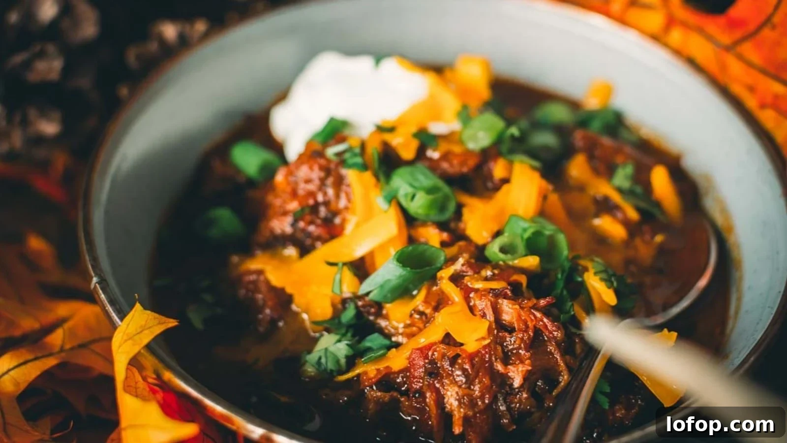 12 Epic Chili Recipes for Game Day Crowd-Pleasing Flavor 8 A bowl of chili topped with shredded cheese, sour cream, and chopped green onions, surrounded by autumn leaves.