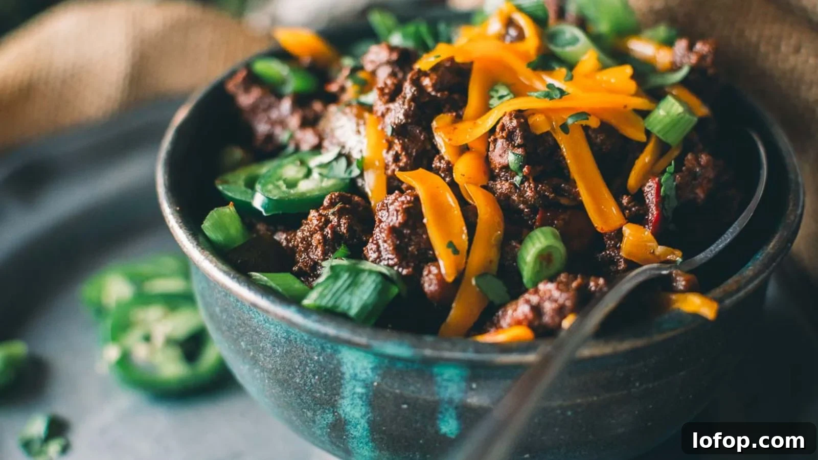12 Epic Chili Recipes for Game Day Crowd-Pleasing Flavor 9 Bowl of chili topped with shredded cheese, sliced jalapeños, and chopped green onions. A spoon rests inside the bowl.