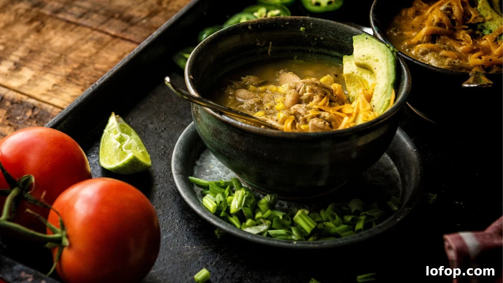 12 Epic Chili Recipes for Game Day Crowd-Pleasing Flavor 10 A bowl of soup with meat, avocado, and green onions on a tray. Nearby are sliced jalapeños, a wedge of lime, whole tomatoes, and a dark wooden surface.