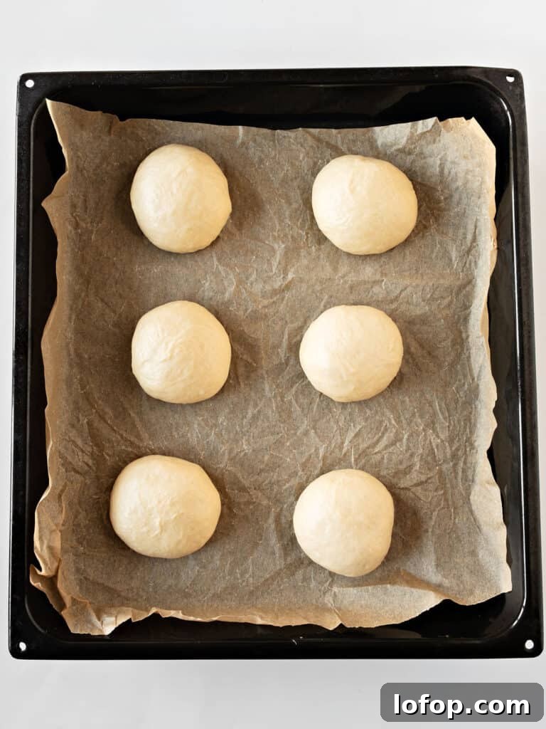 Perfectly Soft Homemade Burger Buns Recipe 8 Parchment paper on a black baking tray with six burger buns before rising.